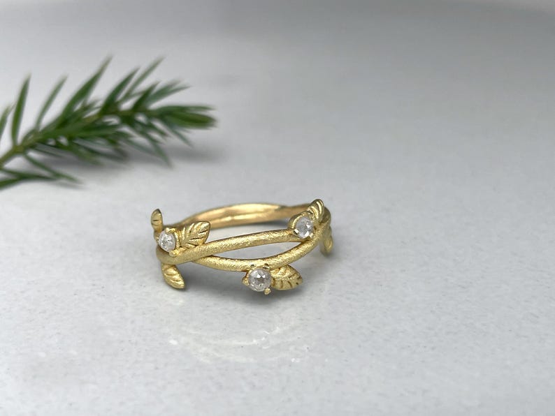 May include: A gold ring with a leaf and vine design. The ring has three small, clear gemstones. The leaves and vines wrap around the band. The ring is on a light grey surface.