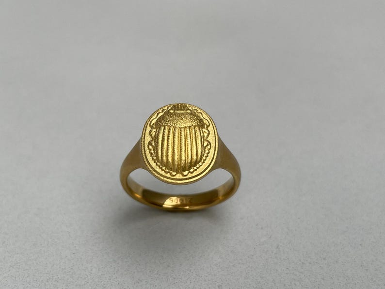 May include: A gold ring featuring a round, raised design of a scarab beetle.