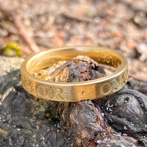 K&ouml;nnte beinhalten: Ein goldener Bandring mit der Inschrift "AMOR" auf der Au&szlig;enseite des Bandes. Die Inschrift ist in einer Serifenschrift und leicht von der Oberfl&auml;che des Bandes erh&ouml;ht. Der Ring liegt auf einem St&uuml;ck Rinde.