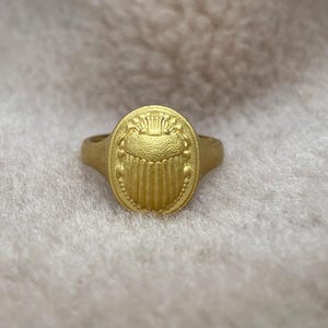 May include: A gold ring featuring an intricate scarab beetle design. The ring is oval in shape and has a textured finish.