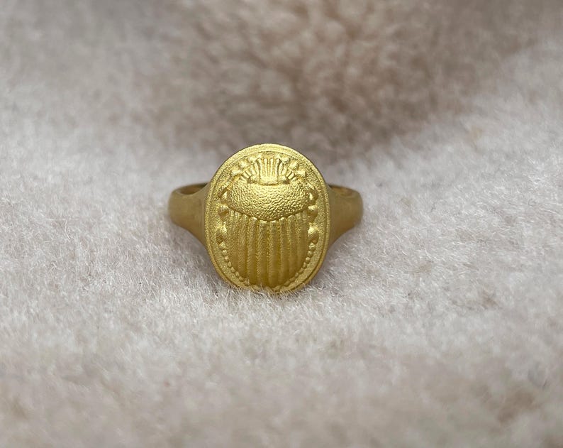 May include: A gold ring featuring an intricate scarab beetle design. The ring is oval in shape and has a textured finish.