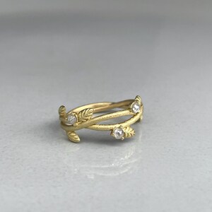 May include: A gold ring with a leaf design. The ring has three clear, round gemstones and a textured finish. The band wraps around the finger with a delicate, nature-inspired aesthetic. The ring is set against a neutral background.
