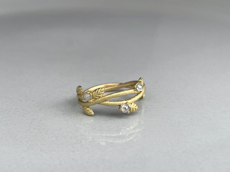 May include: A gold ring with a leaf design. The ring has three clear, round gemstones and a textured finish. The band wraps around the finger with a delicate, nature-inspired aesthetic. The ring is set against a neutral background.