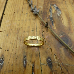 K&ouml;nnte beinhalten: Ein goldener Ring mit der Gravur "VINCIT" auf der Au&szlig;enseite.
