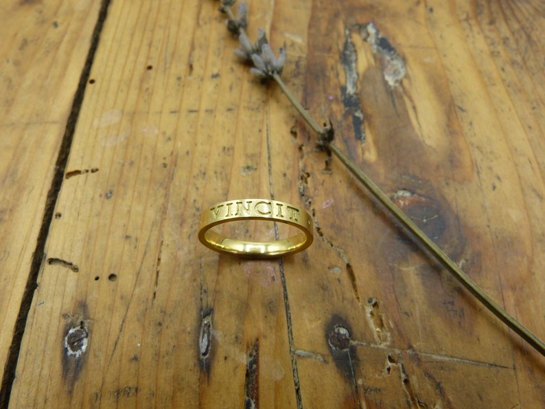 K&ouml;nnte beinhalten: Ein goldener Ring mit der Gravur "VINCIT" auf der Au&szlig;enseite.
