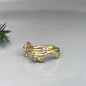 May include: A gold ring with a leaf and vine design. The ring features three small, clear gemstones. The leaves and vines wrap around the band, creating a nature-inspired aesthetic. The ring is set against a neutral background.