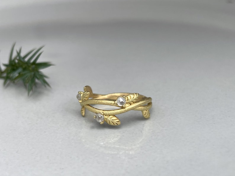 May include: A gold ring with a leaf and vine design. The ring features three small, clear gemstones. The leaves and vines wrap around the band, creating a nature-inspired aesthetic. The ring is set against a neutral background.