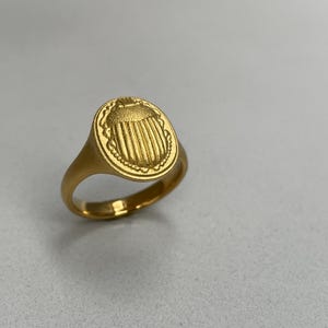 May include: A gold signet ring with an oval shaped top featuring a detailed engraved design of a scarab beetle.