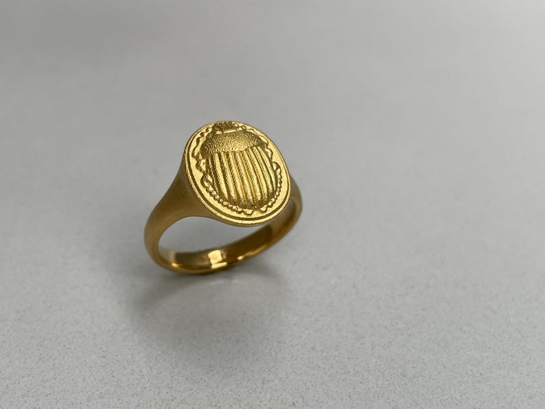 May include: A gold signet ring with an oval shaped top featuring a detailed engraved design of a scarab beetle.