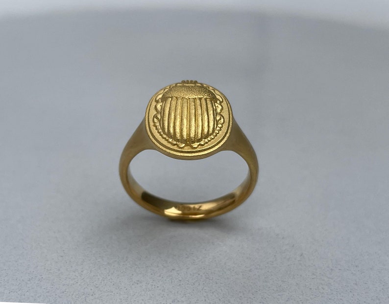 May include: A gold ring featuring a raised scarab beetle design. The ring is oval shaped and has a smooth band.