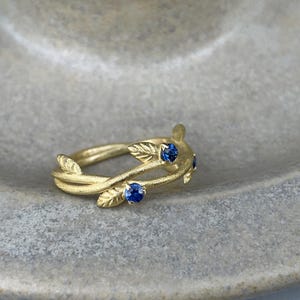 May include: A gold ring with a delicate design of intertwined branches and leaves, accented with four small blue sapphires.