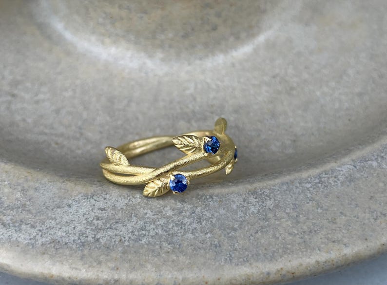 May include: A gold ring with a delicate design of intertwined branches and leaves, accented with four small blue sapphires.