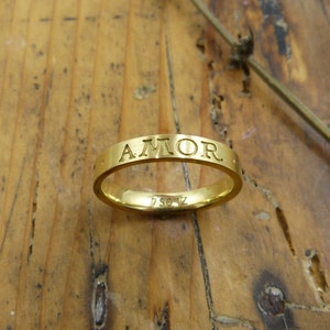 K&ouml;nnte beinhalten: Ein goldener Bandring mit der Gravur "AMOR" auf der Au&szlig;enseite. Der Ring ist innen mit "750 1Z" gekennzeichnet.