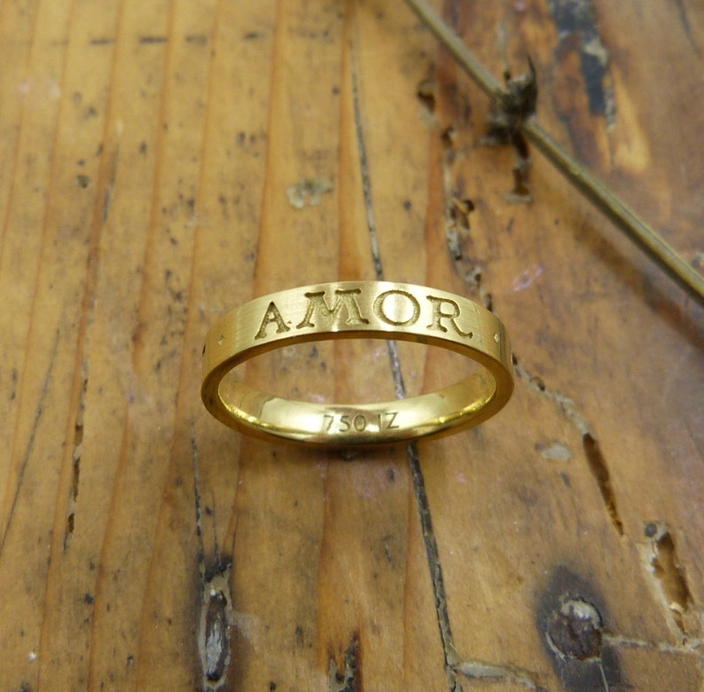 K&ouml;nnte beinhalten: Ein goldener Bandring mit der Gravur "AMOR" auf der Au&szlig;enseite. Der Ring ist innen mit "750 1Z" gekennzeichnet.