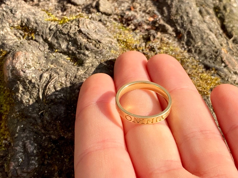 K&ouml;nnte beinhalten: Ein goldener Bandring mit der Gravur "OMNIA" auf der Au&szlig;enseite.