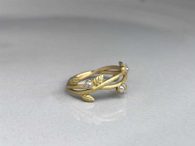 May include: A gold ring with a leaf and vine design. The ring has three small, clear gemstones. The leaves and vines wrap around the band, creating a nature-inspired aesthetic.