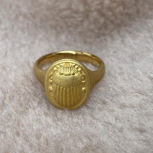 May include: A gold ring with an oval-shaped design featuring a stylised crown and vertical lines. The ring is on a white fluffy surface.