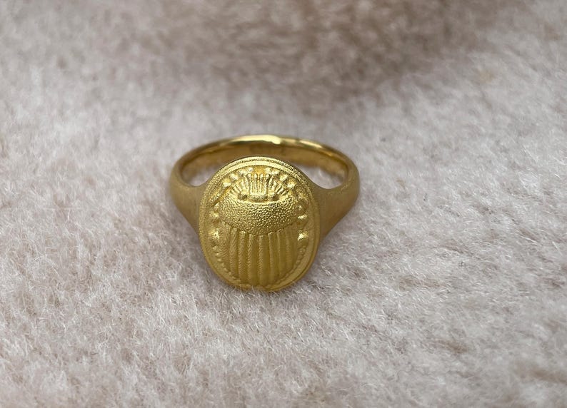 May include: A gold ring with an oval-shaped design featuring a stylised crown and vertical lines. The ring is on a white fluffy surface.