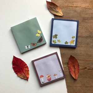 Könnte beinhalten: Drei verschiedene Designs von quadratischen Haftnotizblöcken. Der erste Block hat einen grünen Hintergrund mit einem gelben Schmetterling, einem weißen Schmetterling und einem roten Schmetterling. Der zweite Block hat einen blauen Hintergrund mit grünen Blättern. Der dritte Block hat einen lila Hintergrund mit roten und weißen Pilzen.