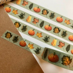 May include: Three rolls of washi tape with a fall theme. The tape features a green background with orange pumpkins, brown pine cones, and yellow and brown leaves.