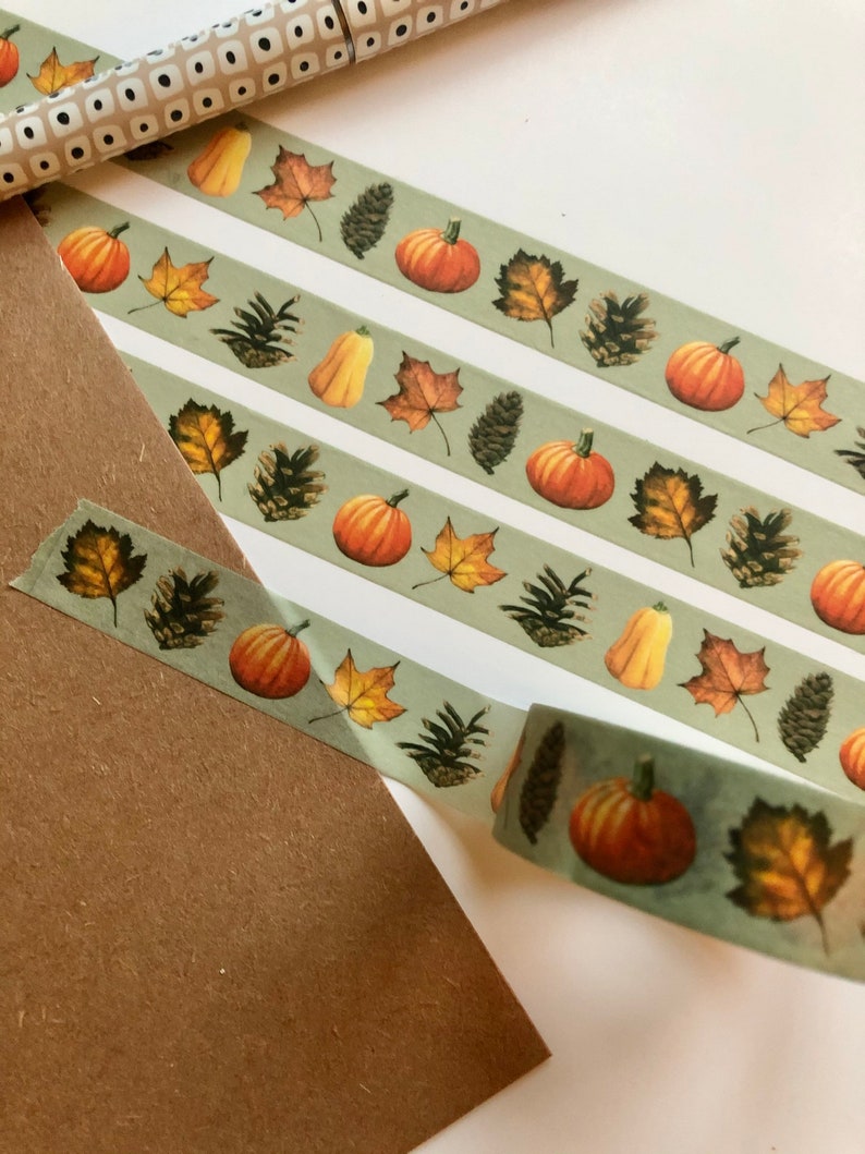 May include: Three rolls of washi tape with a fall theme. The tape features a green background with orange pumpkins, brown pine cones, and yellow and brown leaves.