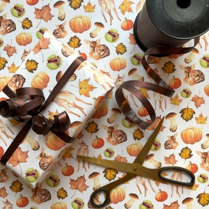 May include: White wrapping paper with a repeating pattern of brown and orange pumpkins, brown mushrooms, brown leaves, and white deer. A brown ribbon is tied around a gift box wrapped in the paper. A pair of gold scissors and a spool of brown ribbon are also in the image.