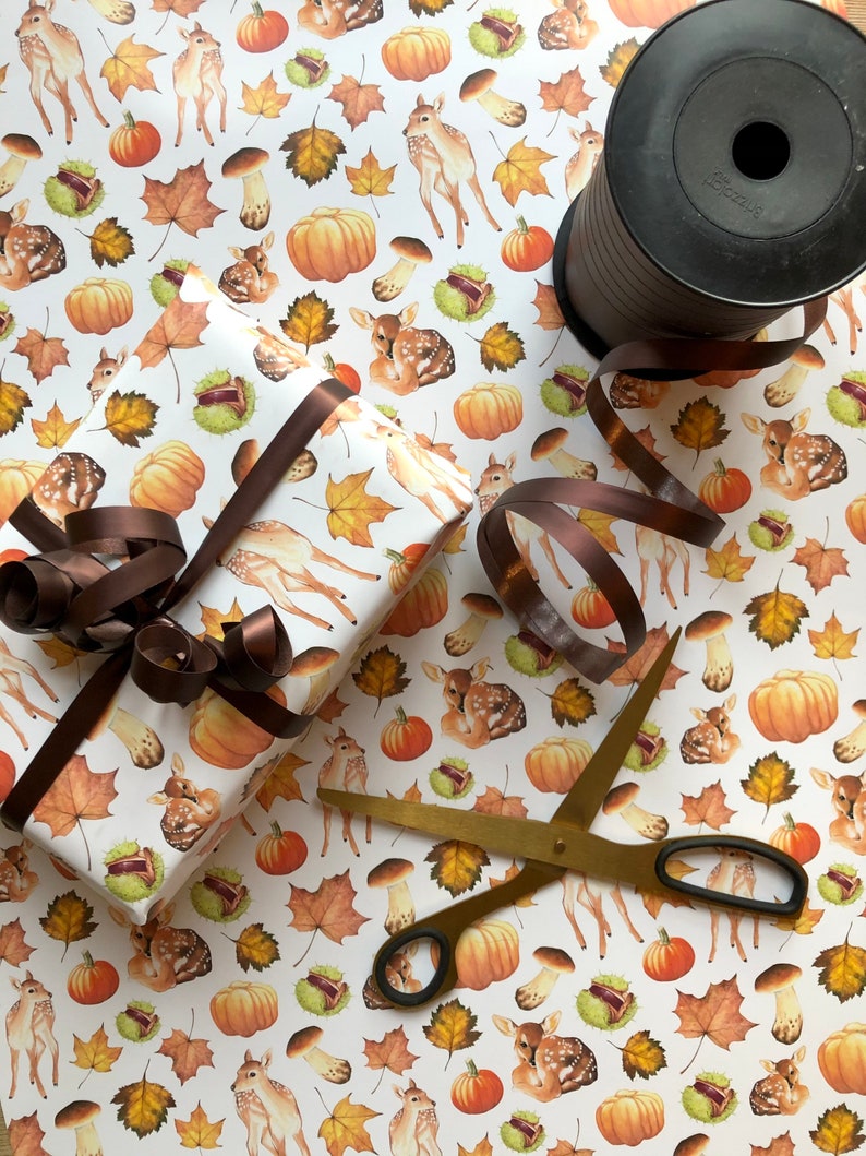 May include: White wrapping paper with a repeating pattern of brown and orange pumpkins, brown mushrooms, brown leaves, and white deer. A brown ribbon is tied around a gift box wrapped in the paper. A pair of gold scissors and a spool of brown ribbon are also in the image.