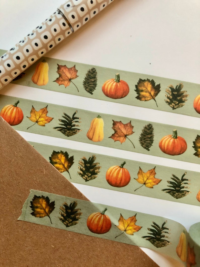 May include: A roll of washi tape with a green background and a repeating pattern of pumpkins, pinecones, and fall leaves.