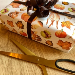 May include: A white gift box wrapped in autumn-themed paper with a brown satin ribbon bow. The paper features illustrations of deer, mushrooms, acorns, and leaves. A pair of gold scissors are lying on a wooden surface.