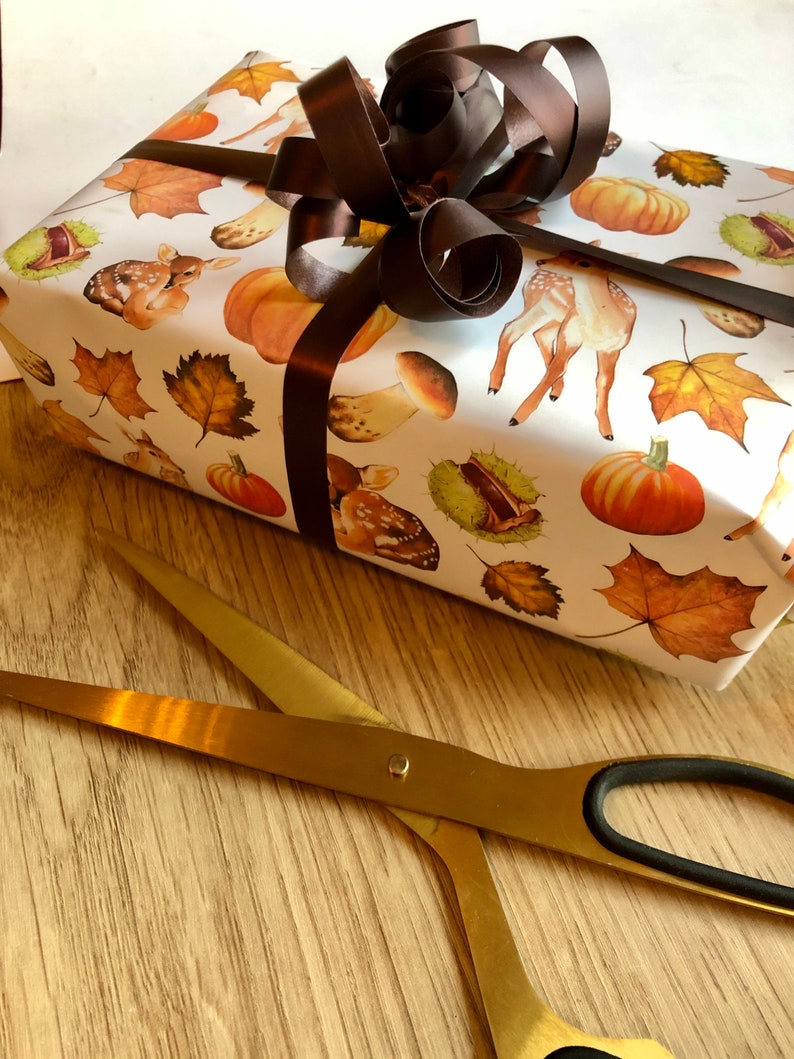 May include: A white gift box wrapped in autumn-themed paper with a brown satin ribbon bow. The paper features illustrations of deer, mushrooms, acorns, and leaves. A pair of gold scissors are lying on a wooden surface.