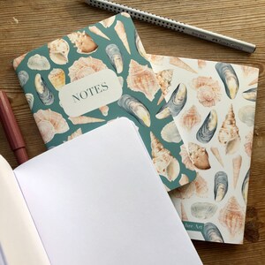 May include: Two notebooks with a teal and white seashell design. The top notebook has the word "NOTES" written on the front. The bottom notebook is open and has blank white pages.