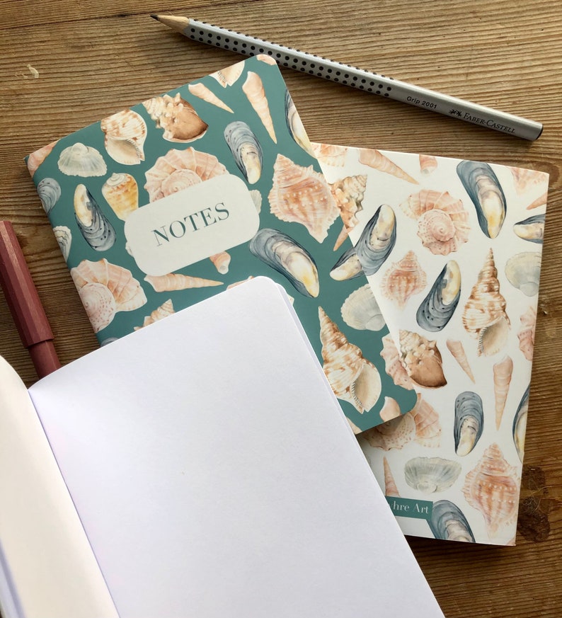 May include: Two notebooks with a teal and white seashell design. The top notebook has the word "NOTES" written on the front. The bottom notebook is open and has blank white pages.