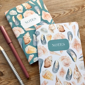 May include: Two teal and white notebooks with a seashell and mussel pattern. The notebooks have the word "NOTES" printed on the front cover.