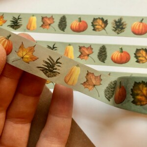 May include: A roll of washi tape with a green background and autumn-themed illustrations. The tape features pumpkins, leaves, pine cones, and gourds.