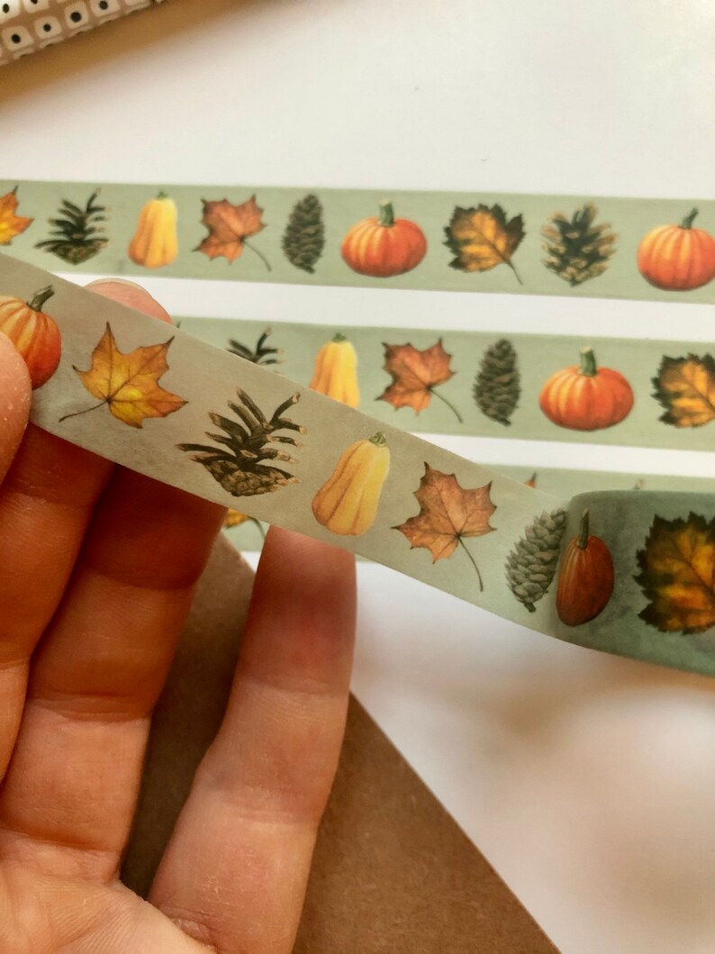 May include: A roll of washi tape with a green background and autumn-themed illustrations. The tape features pumpkins, leaves, pine cones, and gourds.