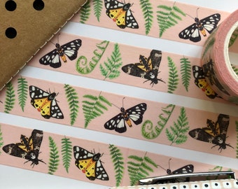 May include: Pink washi tape with a repeating pattern of green ferns and black and yellow butterflies. The tape is rolled up on a spool.
