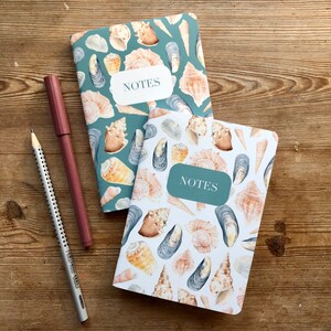 May include: Two small notebooks with a seashell and mussel design. The notebooks are teal and white with the word "NOTES" printed on the front.