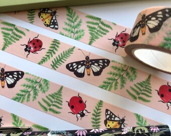 May include: Pink washi tape with a repeating pattern of ladybugs, butterflies, and ferns. The tape is rolled up on a spool and there is a pen with a floral pattern in the foreground.