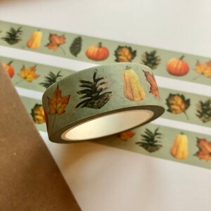 May include: A roll of green washi tape with a fall theme. The tape features illustrations of pumpkins, gourds, pine cones, and leaves in shades of orange, yellow, and brown.