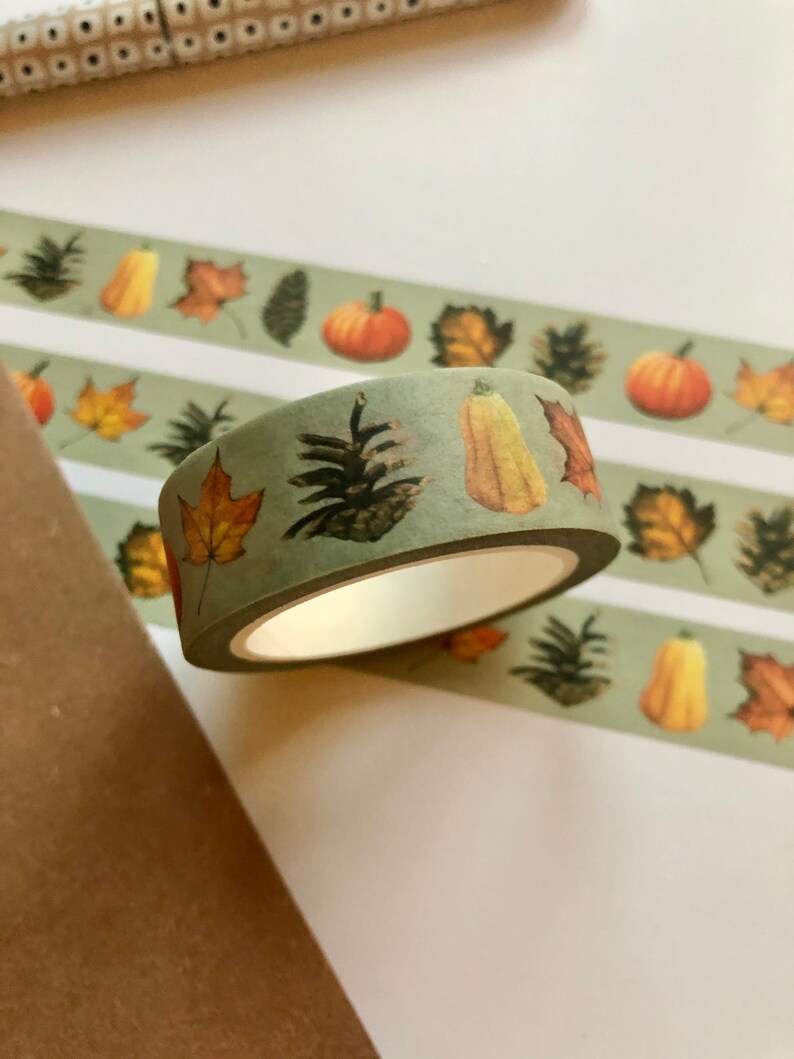 May include: A roll of green washi tape with a fall theme. The tape features illustrations of pumpkins, gourds, pine cones, and leaves in shades of orange, yellow, and brown.