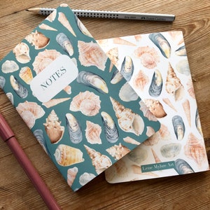 May include: Two notebooks featuring a seashell and mussel design. The top notebook has a teal cover with the word "NOTES" printed on it. The bottom notebook has a white cover with the text "Lene Myhre Art" printed on it.