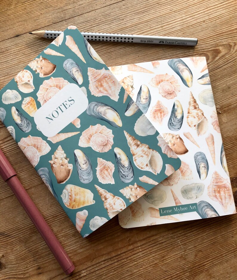 May include: Two notebooks featuring a seashell and mussel design. The top notebook has a teal cover with the word "NOTES" printed on it. The bottom notebook has a white cover with the text "Lene Myhre Art" printed on it.