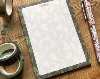 May include: A brown notepad with a white fern pattern on the cover. The notepad has a white interior with the word "NOTES" printed in black at the top. A pen with a floral design is lying next to the notepad.