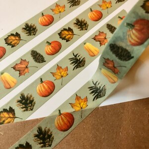 May include: Three rolls of washi tape with a fall theme. The tape has a light green background with a repeating pattern of pumpkins, pine cones, and fall leaves.