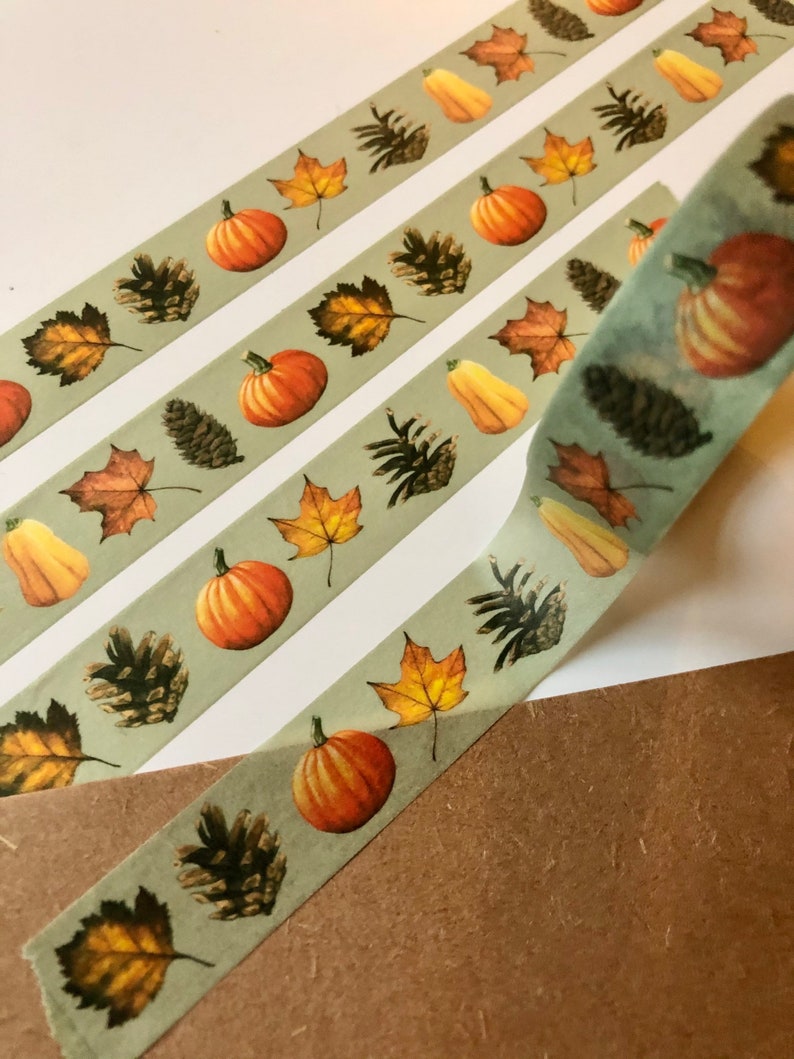 May include: Three rolls of washi tape with a fall theme. The tape has a light green background with a repeating pattern of pumpkins, pine cones, and fall leaves.