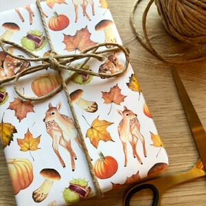 May include: White wrapping paper with a repeating pattern of fall-themed illustrations. The illustrations include deer, pumpkins, mushrooms, leaves, and chestnuts. The paper is tied with twine.