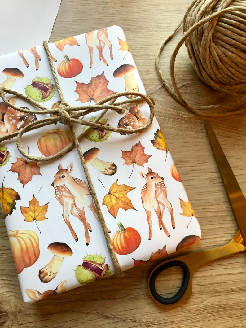 May include: White wrapping paper with a repeating pattern of fall-themed illustrations. The illustrations include deer, pumpkins, mushrooms, leaves, and chestnuts. The paper is tied with twine.