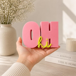 Könnte beinhalten: Ein dreidimensionales, rosafarbenes Blockbuchstaben-Schild mit dem Wort OH in einer fetten, serifenlosen Schrift. Ein kleineres, leuchtend neongelbes Wort hi in Schreibschrift ist an der Vorderseite des Buchstabens H befestigt.