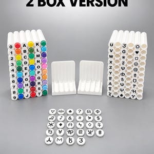 Puede incluir: Dos cajas de almacenamiento de plástico blanco con ranuras cilíndricas, cada una con marcadores coloridos etiquetados con números, letras y símbolos. El texto 2 BOX VERSION está en la parte superior. Una colección de fichas blancas con símbolos negros.