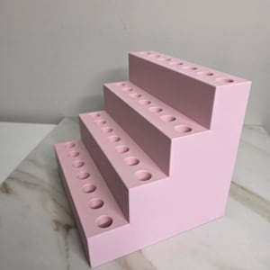 May include: A pink, stepped organizer with multiple circular holes. The organizer has three tiers, each with a row of evenly spaced holes. The item is made of a solid, light pink material and is designed to hold small items.